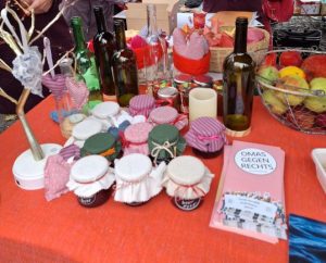 Auf einem roten Tischtuch stehen viele Marmeladengläser mit Stoffhäubchen, dahinter Flaschen auf Holzsockeln, Genähtes und in einem Korb gefilzte Äpfel. Vorne liegen die Flyer der OMAS GEGEN RECHTS aus Hildesheim.