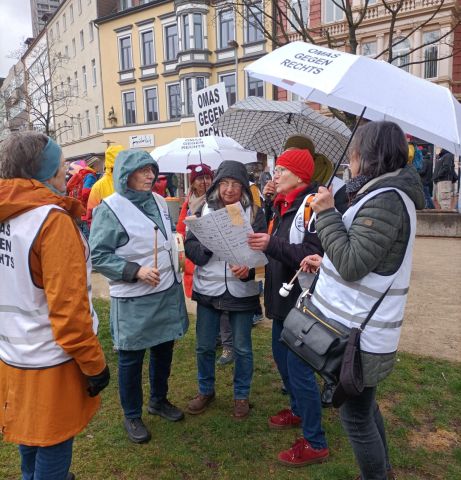 Mehrere Frauen mit weißen Warnwesten und Schirmen stehen auf einer kleinen Wiese und singen.