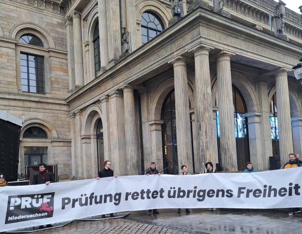 Vor dem Säulenportal der Hannoverschen Oper haben sich mehrere Menschen aufgestellt und halten ein langes Transparent hoch. Darauf ist das Logo von PRÜF Niedersachsen und die ausgeschriebene Erläuterung „Prüfung rettet übrigens Freiheit“.