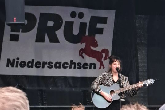 Auf einer Bühne steht eine junge Frau mit Gitarre und singt ins Mikro. Der Bühnenhintergrund ist schwarz und dort hängt ein Schild mit der Aufschrift „PRÜF Niedersachsen“.