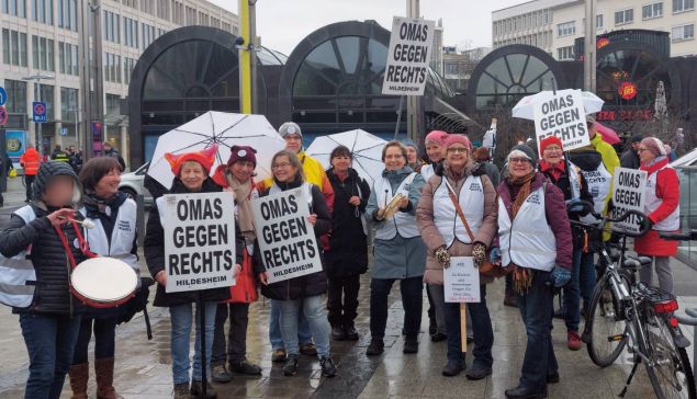 Eine Gruppe von Frauen hat sich vor dem Kröpcke in Hannover aufgestellt. Sie sind warm und regenfest angezogen und haben weiße Westen, Schirme und mehrere Schilder mit der Aufschrift „OMAS GEGEN RECHTS HILDESHEIM“ dabei.
