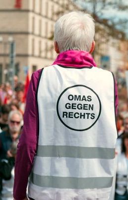 Eine Frau mit kurzen, weißen Haaren und einer magentafarbenen Jacke von hinten. Sie trägt eine weiße Warnweste mit zwei Reflektorstreifen und einem schwarzen Kreis auf dem Rücken, in dem steht "OMAS GEGEN RECHTS".