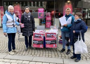Der kleine Infostand mit Plakaten für die PRÜF-Demo in der Mitte, auf dem Tisch Infomaterial, rechts und links stehen jeweils zwei OMAs. Jetzt hält die OMA, die sich eben schnell den Flyer genommen hat, ihn lachend in die Höhe.