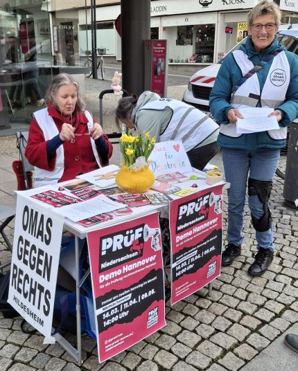 Der rundum mit Plakaten beklebte kleine Infotisch der OMAS GEGEN RECHTS in der Hildesheimer Fußgängerzone. Mitten darauf steht im gelben Übertopf ein Topf mit Narzissen, daneben ein gemaltes Schild, auf dem in bunten Buchstaben steht "Omas für Demokratie". Hinter dem Tisch sitzt eine OMA, rechts daneben steht eine OMA und lacht in die Kamera.