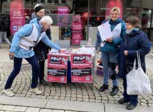 Der kleine Infostand mit Plakaten für die PRÜF-Demo in der Mitte, auf dem Tisch Infomaterial, rechts und links stehen jeweils zwei OMAs. Eine greift sich noch schnell einen Flyer vom Tisch.
