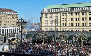 Der Rathausmarkt in Hamburg. Mittig hinten und links mehrstöckige helle Gebäude, zwischen ihnen sieht man die Binnenalster mit Fontäne. Auf dem Platz vor dem Gebäude sind viele Menschen.