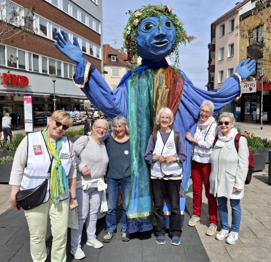 Sechs OMAS GEGEN RECHTS haben sich in einer Reihe aufgestellt und lachen fröhlich. Hinter ihnen steht eine Figur, die doppelt so groß ist wie die Frauen. Sie hat ein blaues Gesicht, Pflanzen als Haare, blaue Hände und trägt ein blaues Gewand, geschmückt mit grünen und braunen Tüchern. Sie hat die Arme ausgebreitet.
