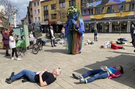 Auf einem hell gepflasterten Platz liegen Menschen in einigen Abständen auf dem Boden. In ihrer Mitte steht eine überlebensgroße Figur, sie hat ihren großen blauen Kopf gesenkt und die blauen Hände in einer betenden Haltung.