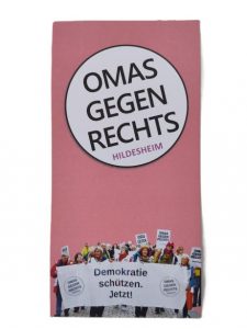 Ein hochformatiger Handzettel: Auf rosa Hintergrund ist das Logo der OMAS GEGEN RECHTS HILDESHEIM, unten sieht man einen Demonstrationszug. Mehrere Frauen tragen ein Banner mit dem Aufdruck "Demokratie schützen. Jetzt!", und aus der Personenmenge ragen Schilder der OMAS GEGEN RECHTS.