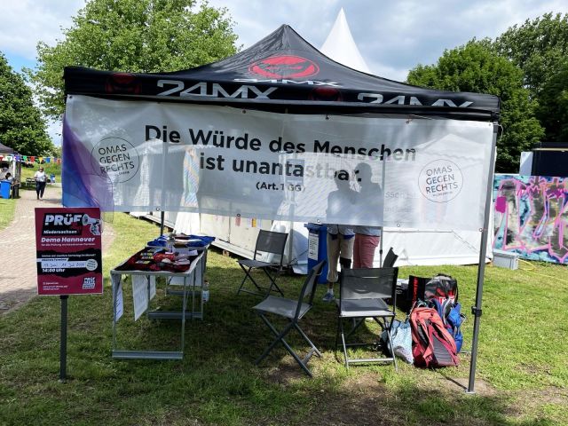 Ein Pavillon, an dem ein Banner befestigt ist mit dem Text „Die Würde des Menschen ist unantastbar. (Art. 1 GG)" Auf dem Banner ist das Logo "OMAS GEGEN RECHTS Hildesheim". Am Pavillon hängt ein Plakat für die PRÜF-Demos in Hannover, unter dem Dach des Pavillons ein langer Tisch mit Infomaterial.