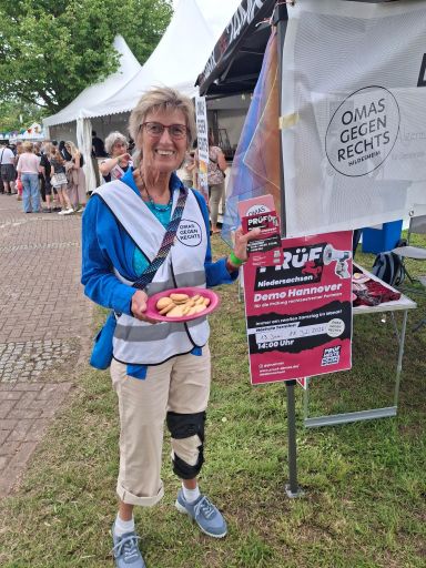 Eine OMA GEGEN RECHTS präsentiert einen Teller mit Keksen und Flyer für die PRÜF-Demo in Hannover. Im Hintergrund sieht man weitere Pavillons entlang des Weges.