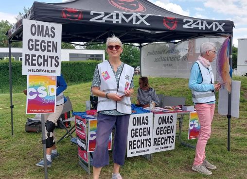 Der Pavillon, geschmückt mit Plakanten für OMAS GEGEN RECHTS und den CSD, davor steht eine OMA und lacht in die Kamera.