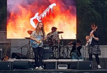 Eine Rockband auf der Bühne: Zwei Personen mit Gitarre und Bass am Bühnenrand, im Hintergrund sieht man das Schlagzeug. Der Bühnenhintergrund zeigt ein Bild mit Flammen, darauf eine weiße E-Gitarre mit dem Bandnamen "Liquid Glass".
