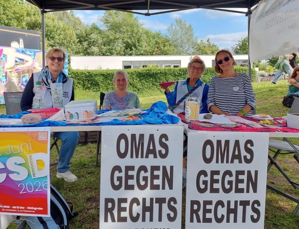 Vier OMAS GEGEN RECHTS sitzen hinter dem Infotisch und lachen in die Kameras. Eine hält einen Teller mit Keksen in die Höhe. Am Tisch hängen Plakate der OMAS GEGEN RECHTS und für den Hildesheimer CSD am 27. Juni 2026.