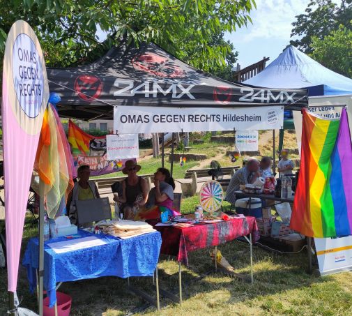 Der Stand der OMAS GEGEN RECHTS Hildesheim auf dem CSD 2026. Unter einem Pavillon mit Banner stehen Tische mit bunten Tischtüchern, darauf verschiedenes Material und ein buntes Glücksrad. Links steht die Beachflag der OMAS, rechts hängt eine Regenbogenfahne.