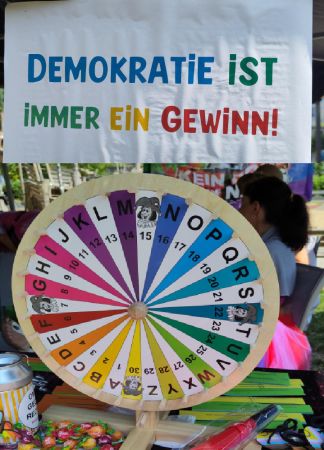Unten ein Glücksrad mit vielen Feldern, abwechselnd weiße und regenbogenbunte Felder. Darüber hängt ein Schild, auf dem in bunten Buchstaben steht: "Demokratie ist immer ein Gewinn!"