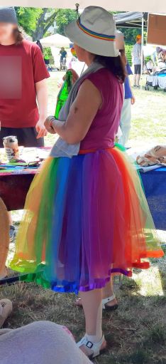 Eine Frau mit Tüllrock in Regenbogenfarben, rotem Shirt und weißem Sonnenhut mit Regenbogen-Band.
