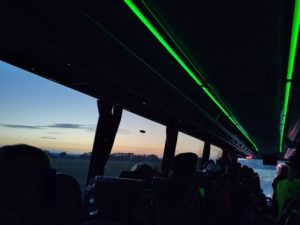 Im Reisebus. Das Innere erscheint schwarz, an der Decke laufen zwei grüne Lichtleisten nach hinten. Draußen sieht man die Morgendämmerung, der Himmel färbt sich allmählich orange.