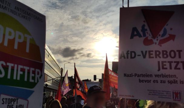 Demozug in der Stadt: Köpfe und Schilder ("AfD-Verbot jetzt") , außerdem Fahnen, die im Gegenlicht rot leuchten.