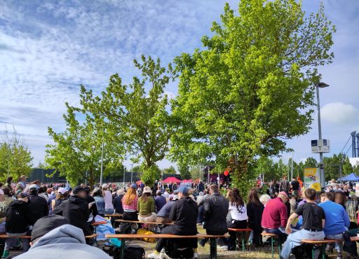 Unter blauem Himmel mit zarten Wölkchen, entlang von grünen Bäumen sitzen auf Bierbänken viele Menschen und schauen zur Bühne, die man im Hintergrund erkennen kann.