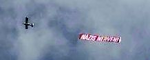 Am zartwolkigen Himmel fliegt ein Flugzeug und zieht ein Transparent hinter sich her. Auf diesem steht Weiß auf Rot "Nazis nerven".