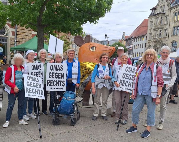 Eine Gruppe OMAS GEGEN RECHTS aus Hildesheim steht um die Maus aus der Kindersendung herum. Sie halten OMA-Schilder hoch.