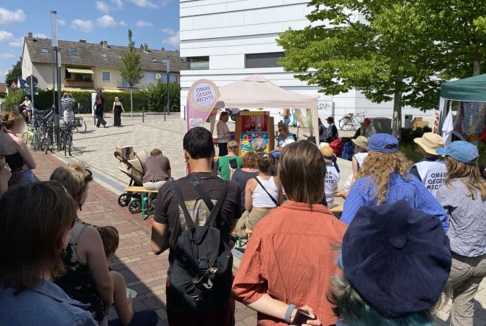 Im Hintergrund der weiße Turm des neuen Mensagebäudes, davor der Pavillon der OMAS GEGEN RECHTS. Dort ist eine Kasperlebühne aufgebaut. Publikum, teils auf Bierbänken sitzend, teils stehend, betrachtet die Vorführung.
