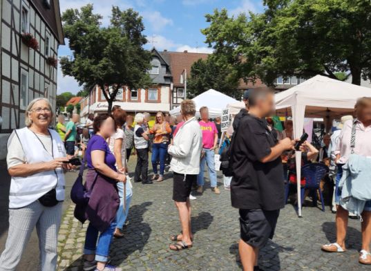 Auf einem gepflasterten Platz vor Fachwerkhäusern stehen viele Menschen im Gespräch. Man erkennt einige OMAS GEGEN RECHTS und rechts zwei Pavillons.