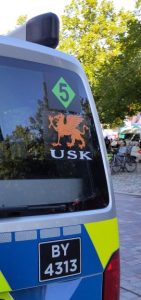 An der Heckscheibe eines Polizeitransporters klebt ein auf der Spitze stehendes grünes Quadrat mit einer 5 darin. Darunter ein orangefarbener Greif mit den Buchstaben USK. Unter der Scheibe ein schwarzes Schild mit "BY 4313".