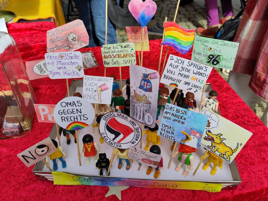 Auf einem roten Tuch steht ein Kasten mit vielen verschiedenen Playmobil-Figuren. Die Playmo-Figuren bilden eine kleine Demonstration und halten viele Schilder hoch. Auf denen steht zum Beispiel "Katzen würden Glatzen kratzen", "Remigriert euch ins Knie" oder "Mir ist kalt und ich wollte ausschlafen! Ihr Sch...Nazis!"