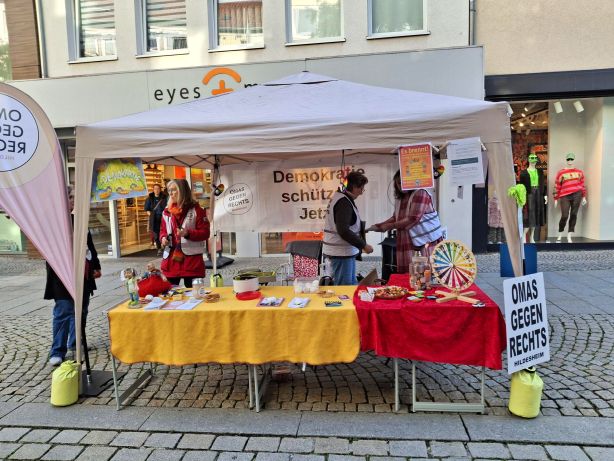Unter einem weißen Pavillon ist in der Fußgängerzone der Stand der OMAS GEGEN RECHTS Hildesheim. Der Pavillon ist mit verschiedenen Plakaten und Bannern geschmückt. Die Tische sind mit gelben und roten Tischtüchern bedeckt, darauf Kekse, Infomaterial und ein buntes Glücksrad. Drei Frauen sind im Stand aktiv.
