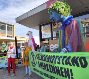 Die überlebensgroße Gaia-Figur mit blauem Gesicht und Pflanzenhaaren steht hinter einem grünen Transparent mit dem Text "Klimanotstand anerkennen" und dem Logo von Extinction Rebellion. Weiter hinten steht eine überlebensgroße Eisbär-Figur mit pinkfarbenem Cape. Links im Bild eine Frau mit weißer Weste und Mikrofon, sie hält eine Rede.
